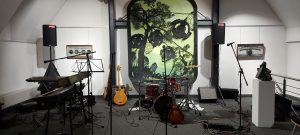 Probenraum, links im Bild Tastenbild, Bildmitte Schlagzeug, Bass, Gitarre, im Hintergrund 3 große hängende Collagen der Künstlerin, rechts eine Skulptur